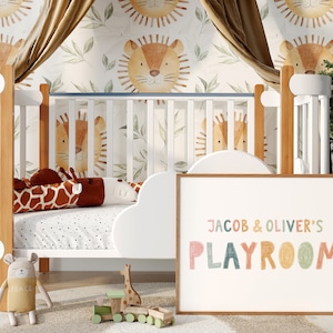 May include: A white crib with a brown wooden frame and a white cloud-shaped bumper. The crib is decorated with a giraffe-shaped pillow and a stuffed animal. The wall behind the crib is covered in a white wallpaper with a pattern of brown lions and green leaves. A framed sign with the text "JACOB & OLIVER'S PLAYROOM" in colorful letters is leaning against the crib.