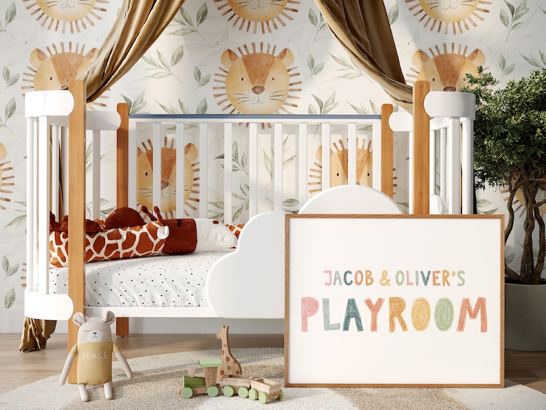 May include: A white crib with a brown wooden frame and a white cloud-shaped bumper. The crib is decorated with a giraffe-shaped pillow and a stuffed animal. The wall behind the crib is covered in a white wallpaper with a pattern of brown lions and green leaves. A framed sign with the text "JACOB & OLIVER'S PLAYROOM" in colorful letters is leaning against the crib.