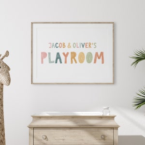 May include: A framed print with the text "Jacob & Oliver's Playroom" in colorful, hand-drawn letters. The print is hanging on a white wall above a wooden dresser.