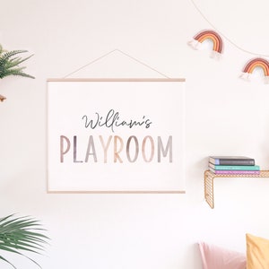 May include: A white canvas print with the text "William's PLAYROOM" in a colorful gradient font. The print is hanging on a white wall with a gold metal shelf above it. The shelf has a potted plant and a stack of books on it. There is a string of rainbow-colored yarn decorations hanging above the print.