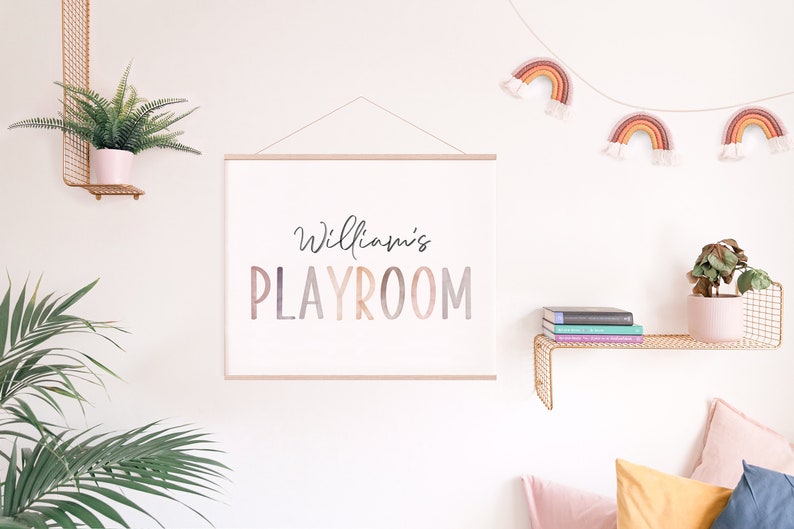 May include: A white canvas print with the text "William's PLAYROOM" in a colorful gradient font. The print is hanging on a white wall with a gold metal shelf above it. The shelf has a potted plant and a stack of books on it. There is a string of rainbow-colored yarn decorations hanging above the print.