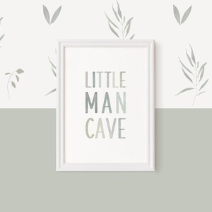 May include: A white framed print with the text "LITTLE MAN CAVE" in a gradient of blue and green colors. The print is hanging on a white wall with a green bottom half. The wall is decorated with small gray leaf designs.
