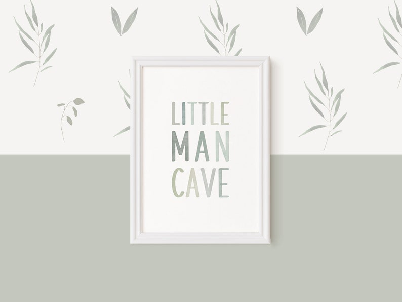 May include: A white framed print with the text "LITTLE MAN CAVE" in a gradient of blue and green colors. The print is hanging on a white wall with a green bottom half. The wall is decorated with small gray leaf designs.