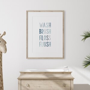 May include: A framed print with the words "WASH BRUSH FLOSS FLUSH" in a gradient of blue colors. The print is hanging on a white wall above a wooden dresser.