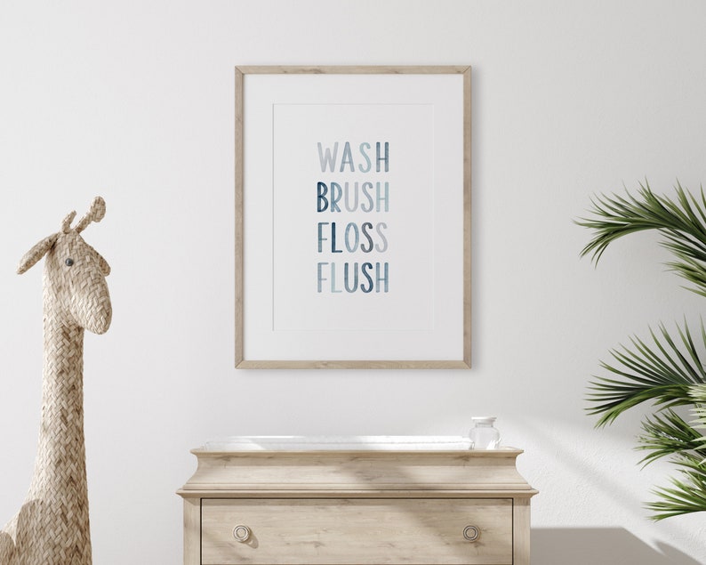 May include: A framed print with the words "WASH BRUSH FLOSS FLUSH" in a gradient of blue colors. The print is hanging on a white wall above a wooden dresser.