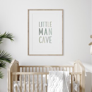 May include: A framed print with the text "LITTLE MAN CAVE" in a gradient of blue and green colors. The print is hanging on a white wall above a wooden crib.