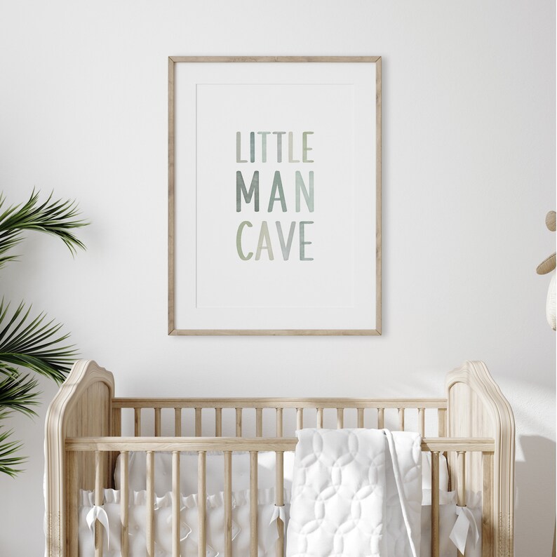 May include: A framed print with the text "LITTLE MAN CAVE" in a gradient of blue and green colors. The print is hanging on a white wall above a wooden crib.