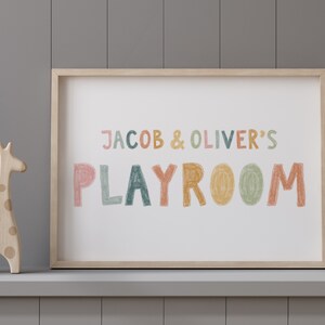 May include: A framed print with the text "JACOB & OLIVER'S PLAYROOM" in colorful, hand-drawn letters. The letters are in shades of pink, orange, yellow, green, and blue. The print is on a white background and is framed in a light wood frame. A wooden giraffe toy is in the foreground.