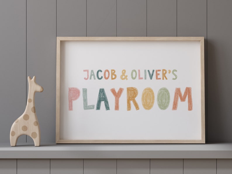 May include: A framed print with the text "JACOB & OLIVER'S PLAYROOM" in colorful, hand-drawn letters. The letters are in shades of pink, orange, yellow, green, and blue. The print is on a white background and is framed in a light wood frame. A wooden giraffe toy is in the foreground.
