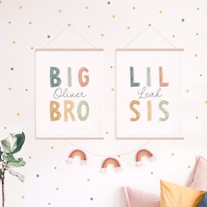 May include: Two framed prints with colorful text that reads "BIG Oliver BRO" and "LIL Leah SIS". The prints are hung on a white wall with a colorful confetti pattern.