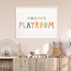 May include: A white framed print with the text "BENJAMIN'S PLAYROOM" in colorful, hand-drawn letters. The print is hanging on a light pink wall above a wooden shelf with various children's toys, including a rainbow abacus, a wooden giraffe, and a toy train.