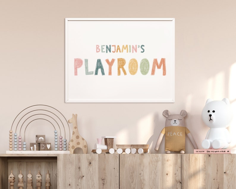 May include: A white framed print with the text "BENJAMIN'S PLAYROOM" in colorful, hand-drawn letters. The print is hanging on a light pink wall above a wooden shelf with various children's toys, including a rainbow abacus, a wooden giraffe, and a toy train.