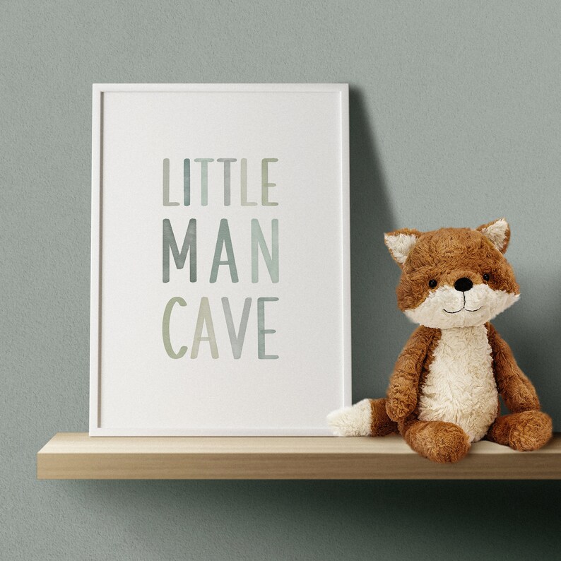 May include: A white framed print with the text "LITTLE MAN CAVE" in a watercolor style. A brown plush fox toy is sitting on a wooden shelf below the print.
