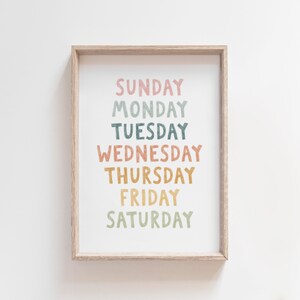 May include: A framed print with the days of the week written in a colorful, hand-drawn style. The words are arranged vertically, with "Sunday" at the top and "Saturday" at the bottom. The colors used are pink, green, blue, orange, and yellow.