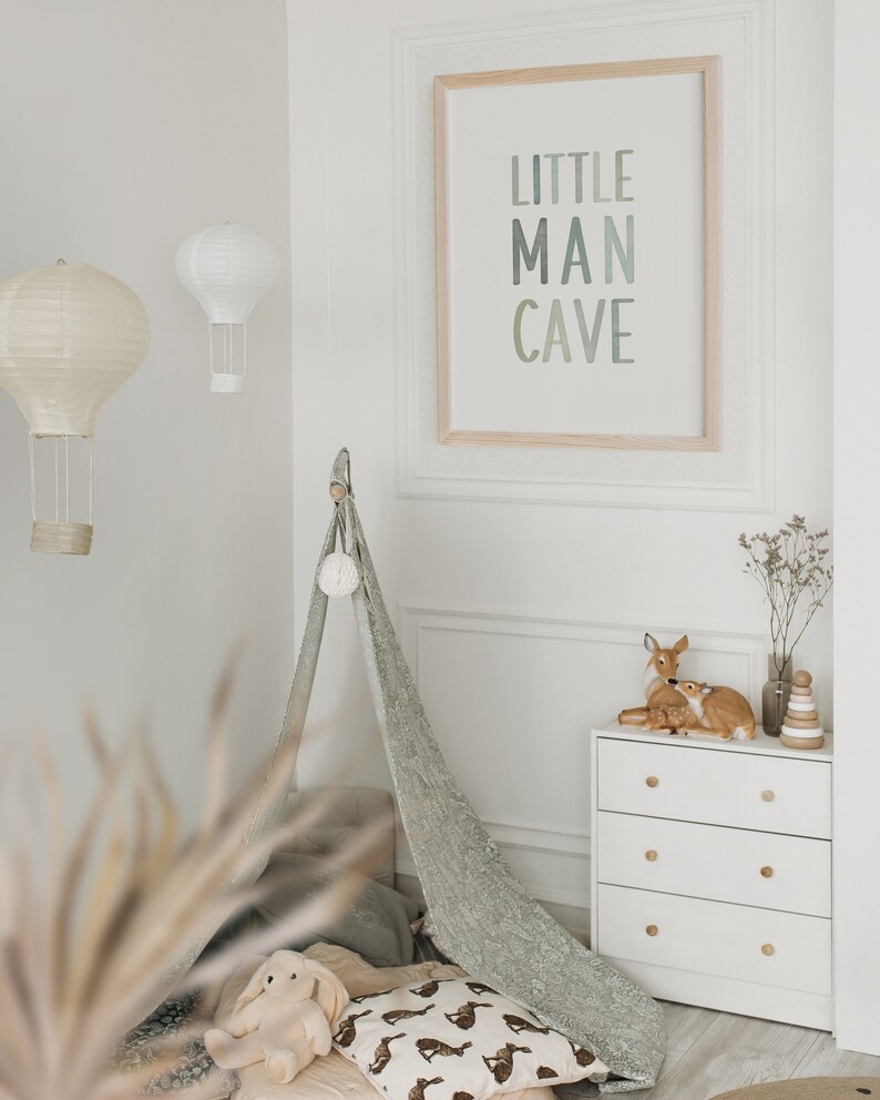 May include: A white dresser with three drawers and wooden knobs. A teepee tent with a white pom-pom on top is in the background. A framed print with the text "LITTLE MAN CAVE" is on the wall. Two small deer figurines are on top of the dresser.