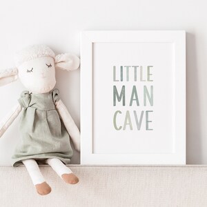 May include: A white framed print with the text "LITTLE MAN CAVE" in a gradient of green and blue colors. A white stuffed animal with a green dress sits on a beige surface in front of the print.