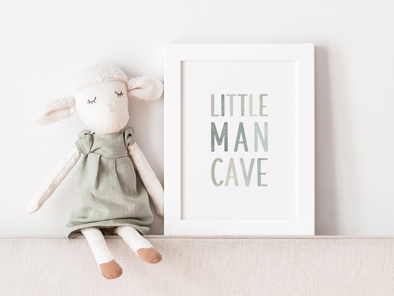 May include: A white framed print with the text "LITTLE MAN CAVE" in a gradient of green and blue colors. A white stuffed animal with a green dress sits on a beige surface in front of the print.