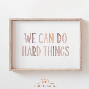 Puede incluir: Una impresión enmarcada con el texto "WE CAN DO HARD THINGS" en un estilo acuarela con tonos de rosa, morado y marrón. La impresión está en un marco de madera claro y tiene un fondo blanco.