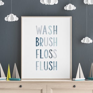 May include: A white framed print with the words "WASH BRUSH FLOSS FLUSH" in blue, gray, and white text on a white background. The print is hanging on a dark gray wall with white paper clouds hanging from the ceiling.