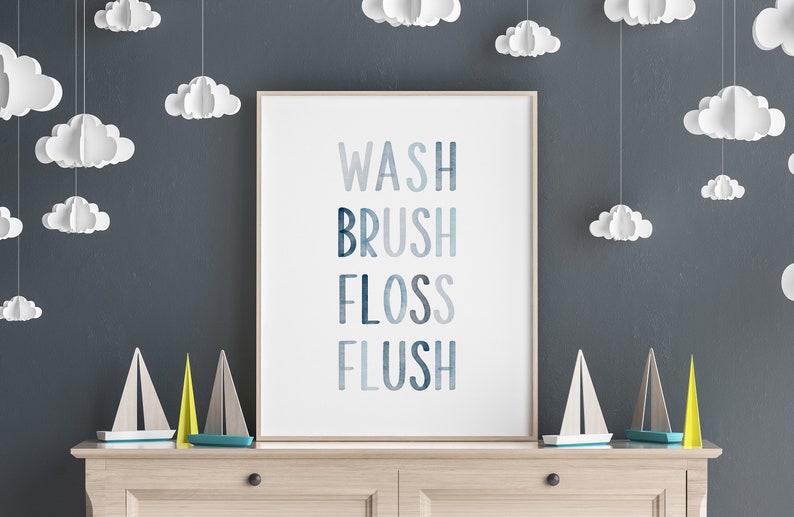 May include: A white framed print with the words "WASH BRUSH FLOSS FLUSH" in blue, gray, and white text on a white background. The print is hanging on a dark gray wall with white paper clouds hanging from the ceiling.