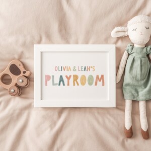 May include: A white framed print with the text "Olivia & Leah's Playroom" in colorful letters. The print is on a beige surface with a wooden toy elephant and a stuffed white lamb doll.