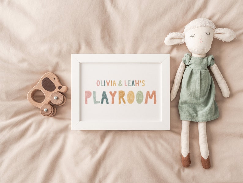 May include: A white framed print with the text "Olivia & Leah's Playroom" in colorful letters. The print is on a beige surface with a wooden toy elephant and a stuffed white lamb doll.