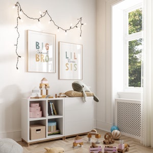 May include: A white bookshelf with two shelves and four cubbies. The shelves are filled with books and toys. There are two framed prints on the wall above the bookshelf. The prints say "Big Bro Samuel" and "Lil Sis Reese".