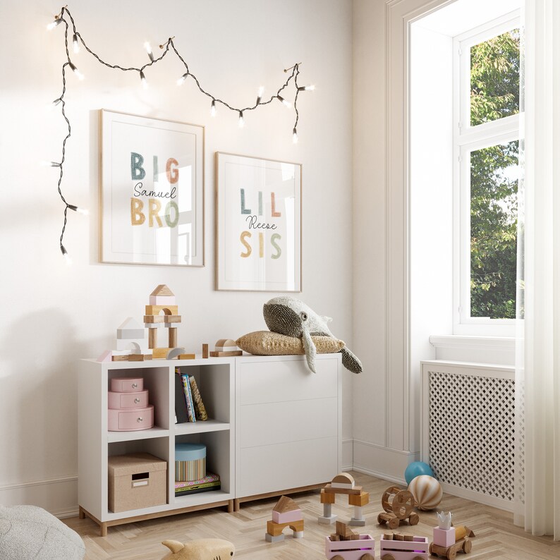 May include: A white bookshelf with two shelves and four cubbies. The shelves are filled with books and toys. There are two framed prints on the wall above the bookshelf. The prints say "Big Bro Samuel" and "Lil Sis Reese".