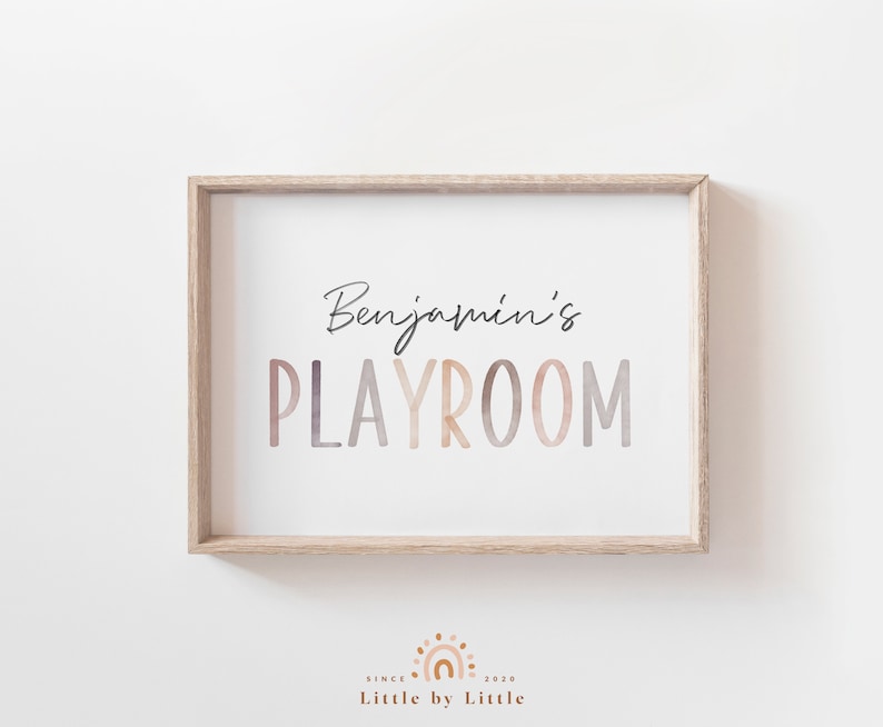 May include: A framed print with the text "Benjamin's Playroom" in a watercolor-style font with a gradient of pink, orange, and brown colors. The frame is a light wood color.