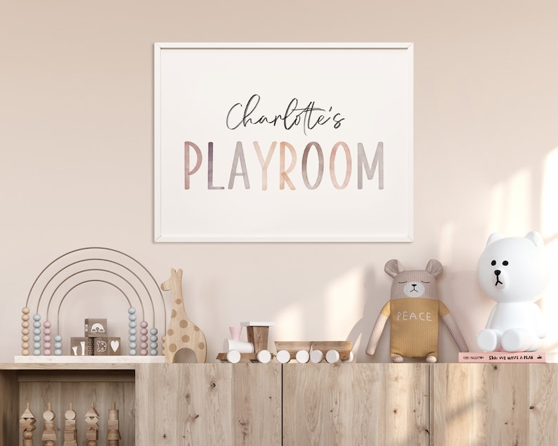 May include: A white framed print with the text "Charlotte's Playroom" in a colorful watercolor font. The print is hanging on a light pink wall above a wooden shelf with a rainbow abacus, wooden giraffe, and other toys.