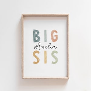 May include: A framed print with the text "BIG SIS" in colorful, hand-drawn letters. The name "Amelia" is written in cursive below the letters.
