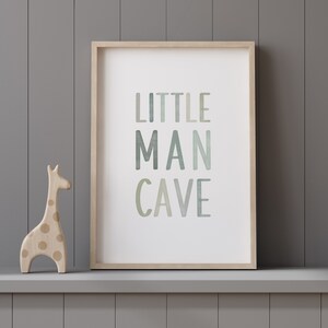 May include: A framed print with the text "LITTLE MAN CAVE" in a watercolor-style font with shades of green and blue. A wooden giraffe figurine with brown spots is in the foreground.