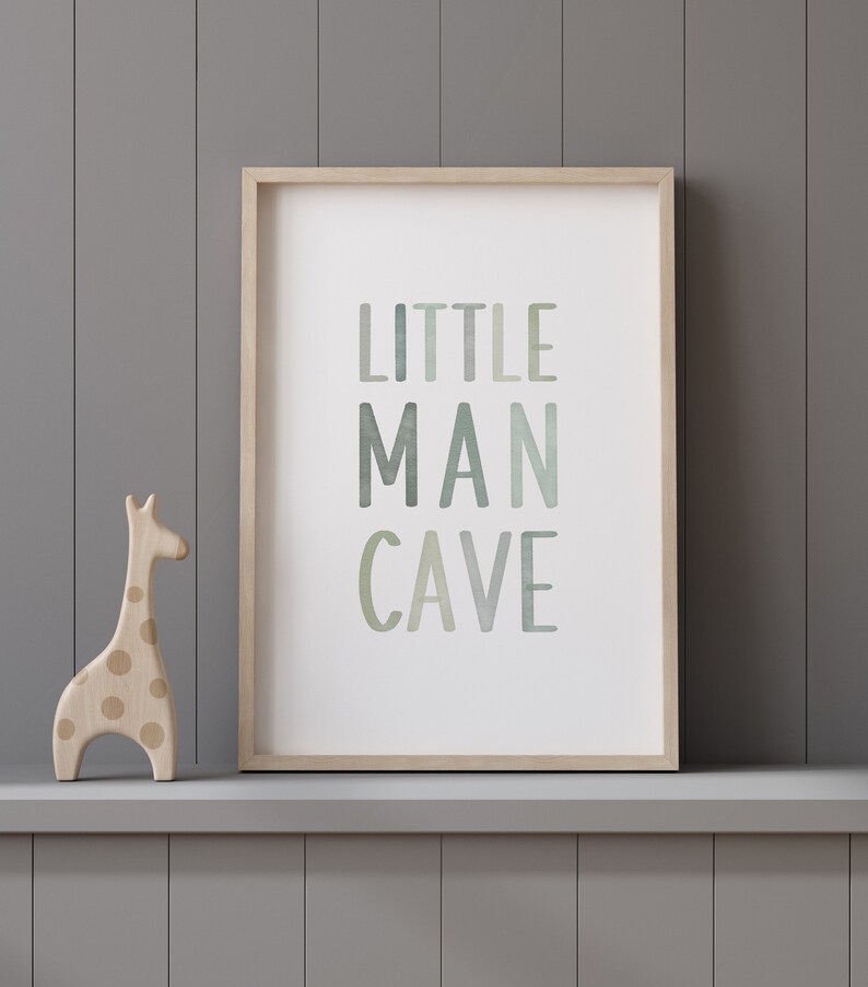 May include: A framed print with the text "LITTLE MAN CAVE" in a watercolor-style font with shades of green and blue. A wooden giraffe figurine with brown spots is in the foreground.