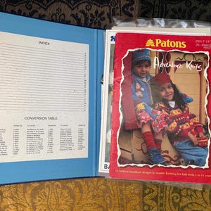 May include: An open blue binder with an index and conversion table, next to a red Patons "Adventure Knits" booklet. The booklet shows two children in colourful knitwear. The booklet's cover has the text "PKN F 317" and "20-30cm".