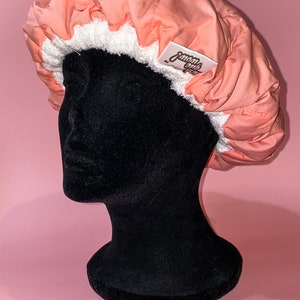 May include: A peach-colored shower cap with a white, fluffy terry cloth lining. The cap has a label that reads "Smooth Girl". The cap is displayed on a black mannequin head against a pink background.