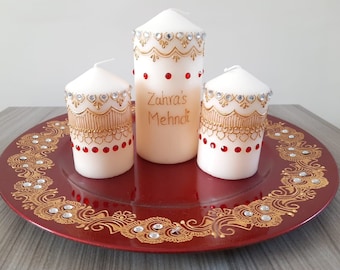 Puede incluir: Tres velas blancas decoradas con diseños de henna dorados y pedrería roja y transparente. Las velas están sobre un plato rojo con un diseño de henna dorado y pedrería. La vela del medio tiene el texto "Zuhra's Mehndi" escrito en ella.