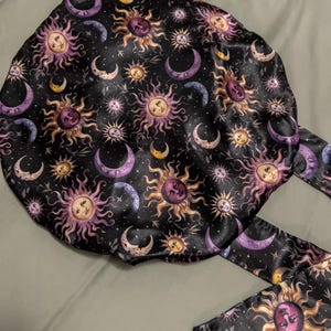 May include: A black satin sleep cap with a repeating pattern of gold and purple suns and moons. The cap has a long tie that is the same fabric as the cap.