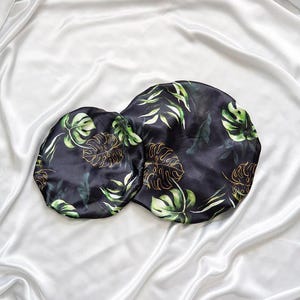 May include: Two black satin hair bonnets with a tropical leaf print in shades of green and gold. The bonnets are on a white satin background.