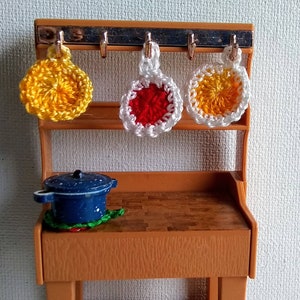 Puede incluir: Un juego de cocina de madera en miniatura con una olla azul en la encimera y tres agarraderas de olla de ganchillo colgadas de ganchos. Las agarraderas de olla son amarillas, rojas y amarillas y blancas.