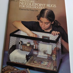 May include: A person holds a book titled "Susan McBaine Miniature Needlepoint Rugs for Dollhouses". The book cover shows a miniature dollhouse with a bed, fireplace, and rugs. The cover is brown with white text.