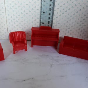 May include: A collection of four small, red plastic furniture items: a sofa, two armchairs, and a piano. The furniture is set on a white surface, with a ruler in the background. These pieces are likely for a doll's house or similar toy.