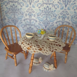 Può includere: Un set da pranzo in miniatura per case di bambole con due sedie in legno, un tavolo con una tovaglia all'uncinetto, due ciotole, un vaso con fiori e due gatti che dormono sotto il tavolo.