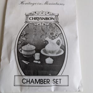 Op de afbeelding: Een witte papieren verpakking met het opschrift "Heritage in Miniatures" en een zwart-wit illustratie van een kamer set. De illustratie bevat een kan, een kom, een kopje en andere miniatuur items. De verpakking is gelabeld "CHAMBER SET".