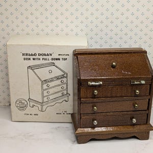 May include: Miniature wooden desk with pull-down top, featuring three drawers and brass knobs. The desk is brown and sits next to its original box, which reads "HELLO DOLLY" and "DESK WITH PULL-DOWN TOP."