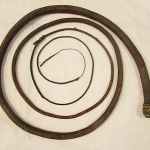 May include: A brown leather whip with a braided handle and a woven end. The whip is coiled in a spiral, with a smaller, thinner whip and a black cord inside the coil. The whip is a dark brown color.