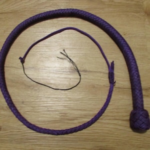 May include: A purple whip with a braided handle and a long, thin lash. The whip is made of a woven material and lies on a wooden surface. The lash is black and appears to be made of several strands.