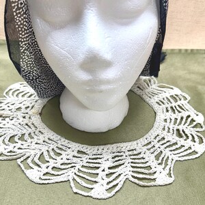 May include: A white, crocheted collar with a scalloped edge is displayed on a white mannequin head. A black and white patterned scarf is draped over the head. The collar is made of a delicate, open weave.