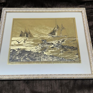 Op de afbeelding: Ingelijste nautische kunstafdruk met een goud-zwarte illustratie van zeilboten op een woelige zee met zeemeeuwen. De afdruk is gevat in een witte passe-partout en een decoratieve witte lijst.