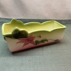 May include: A rectangular ceramic planter with a scalloped rim, featuring a gradient of pale yellow to white and a pink base. The exterior is decorated with a painted design of a large pink flower with green leaves and a small bud on the side.