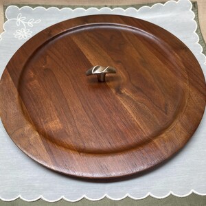 Peut inclure: Un plateau de service rond en bois avec une finition marron foncé. Le plateau présente un bord surélevé et une poignée décorative en métal au centre. Le plateau est posé sur un tissu blanc et vert à bords festonnés.